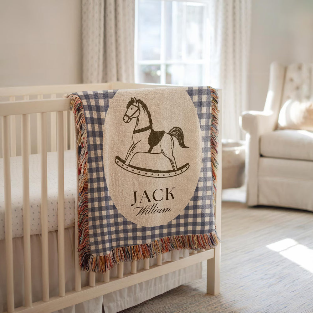 A personalized baby blanket draped over a white wooden crib in a softly lit nursery. The blanket has a navy and beige gingham border, a central beige oval patch with a black rocking horse illustration, and the names 'JACK' and 'William' printed below.