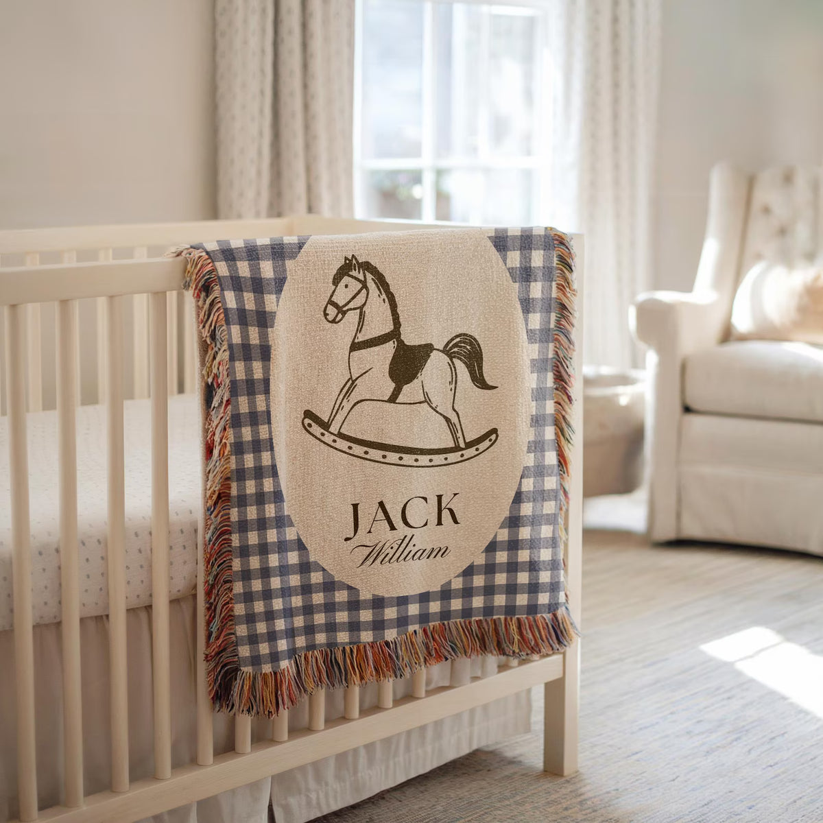 A personalized baby blanket draped over a white wooden crib in a softly lit nursery. The blanket has a navy and beige gingham border, a central beige oval patch with a black rocking horse illustration, and the names 'JACK' and 'William' printed below.