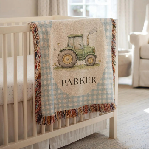 A personalized baby crib blanket draped over the side of a white wooden crib in a softly lit nursery. The blanket features a light blue and white gingham border, a central cream panel with a green tractor illustration and the name 'PARKER' in black text, and multicolored fringe along the edges.